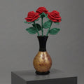Three red knitted roses with green stems and leaves arranged in a small black vase with a gold-toned body, displayed on a gray pedestal against a gray studio background.