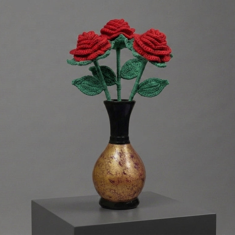 Three red knitted roses with green stems and leaves arranged in a small black vase with a gold-toned body, displayed on a gray pedestal against a gray studio background.