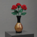 Three red knitted roses with green stems and leaves arranged in a small black vase with a gold-toned body, displayed on a gray pedestal against a gray studio background.