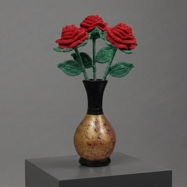 Three red knitted roses with green stems and leaves arranged in a small black vase with a gold-toned body, displayed on a gray pedestal against a gray studio background.